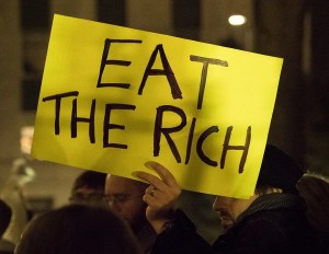A hand-lettered cardboard sign that reads "Eat the Rich" in black capitals on a bright yellow background. The sign is being held by a man in the middle of a groups of protesters.