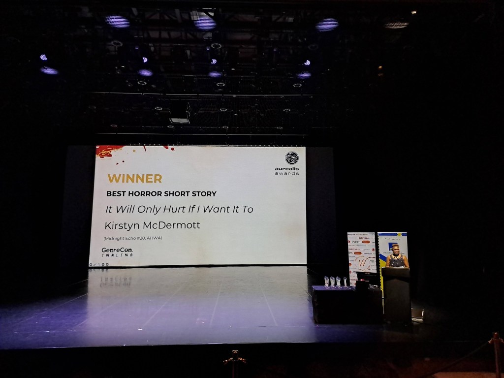 Author extraordinaire and host of the Aurealis Awards 2025, Eugen Bacon, stands at a podium on a large open stage. Behind her is a huge screen announcing the winner of the Best Horror Short Story - "It Will Only Hurt If I Want It To" by Kirstyn McDermott. A table of sharp and pointy Aurealis Awards stand ready to be deployed.