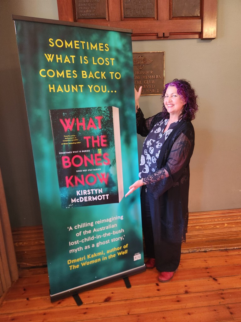 The author, Kirstyn McDermott, standing next to a massive banner promoting her new novel, What the Bones Know. Above a cover image of the book, is yellow text that reads "Sometimes what is lost comes back to haunt you." Below is a quote from Dmetri, Kakmi, author of The Woman in the Well, which reads: "A chilling reimagining of the Australian lost-child-in-the-bush myth as a ghost story."
Photo by Jason Nahrung