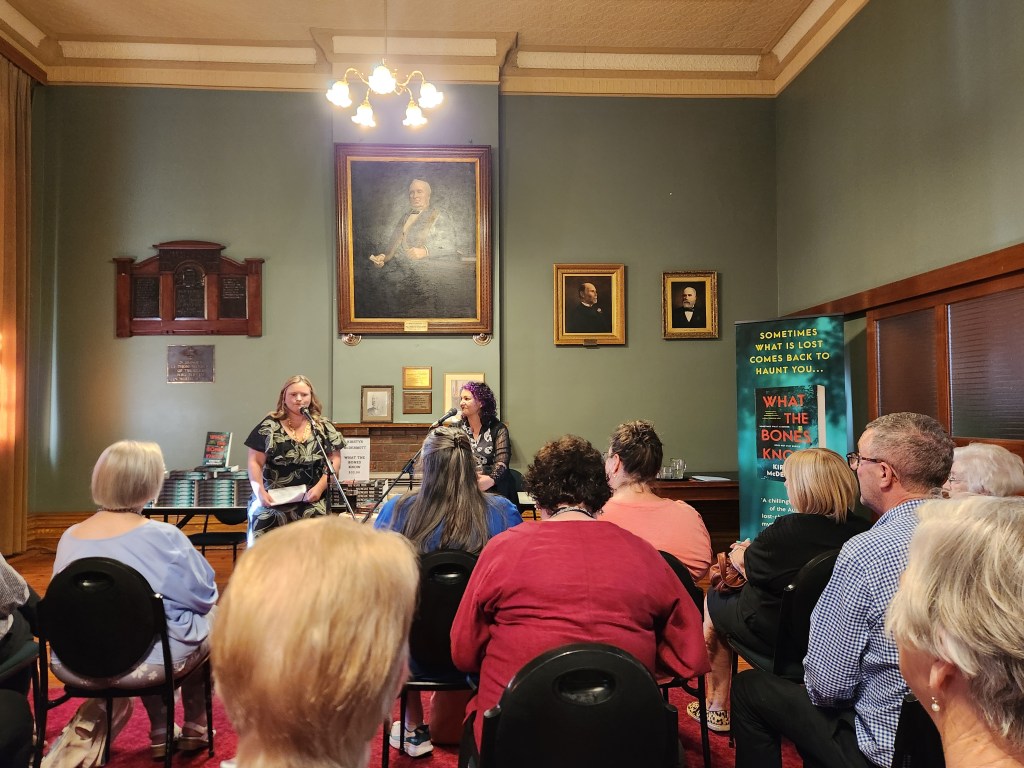 Kirstyn McDermott in conversation with Renee from the Collins Booksellers team. The two sit on chairs at the front of a large, high-ceiling room. The photo has been taken from among the audience.
Photo by Jason Nahrung