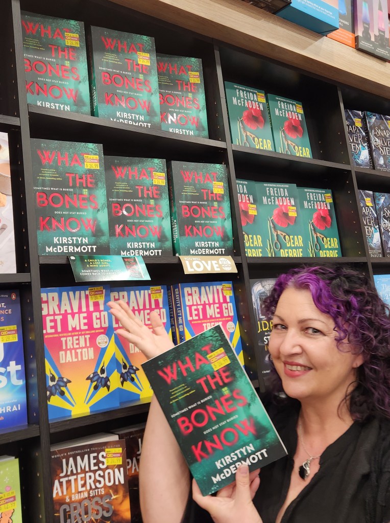The author, Kirstyn McDermott, standing in front of a display shelf of her newly released novel, What the Bones Know. She is also holding a copy of the book and wearing a very, very happy author smile!