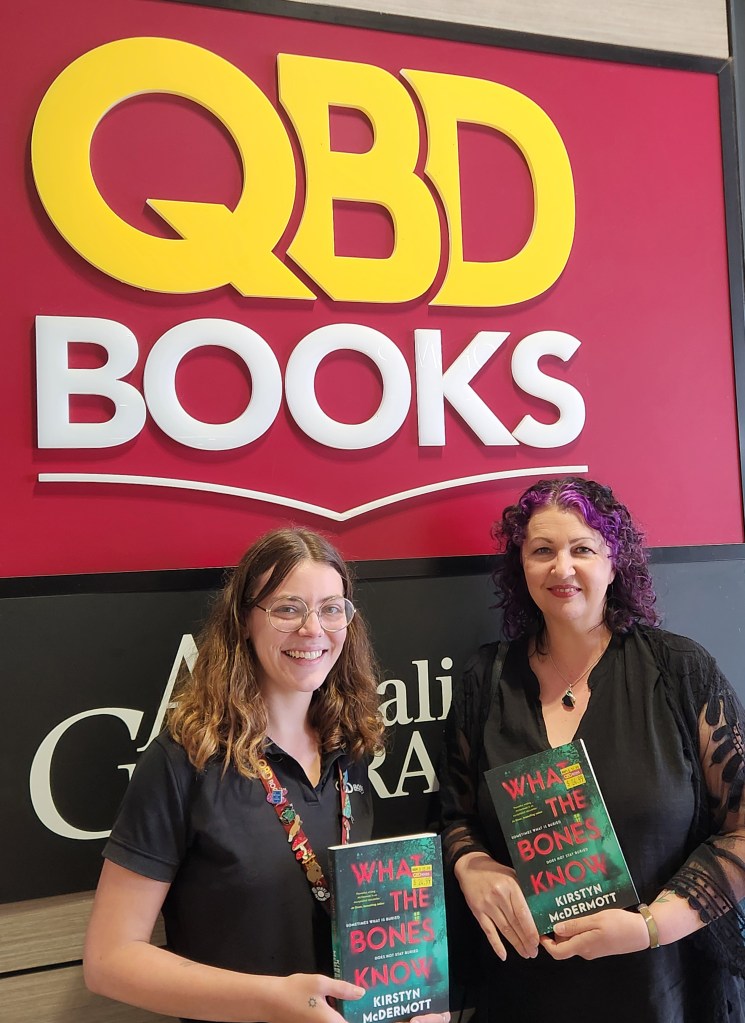 Kirstyn McDermott and Elli from QBD Traralgon, standing in front off the store sign holding copies of What the Bones Know.